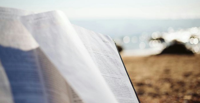 Öppen bok i förgrunden, strand och hav i bakgrunden.