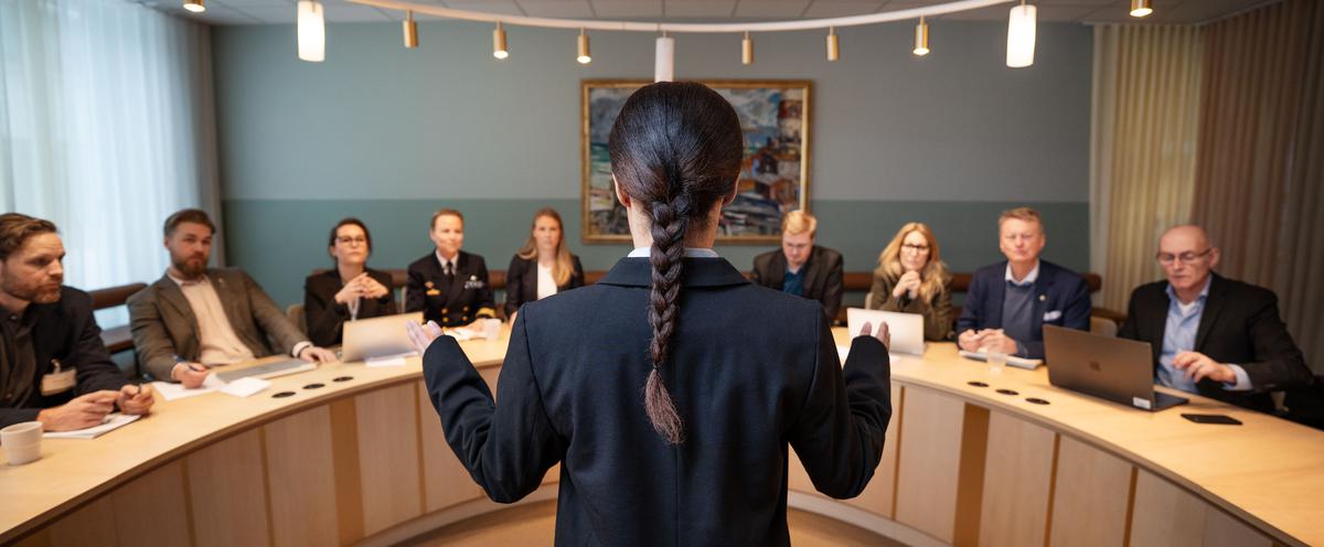 Kvinnlig student med ryggen mot kameran inför en panel av konferensdeltagare.